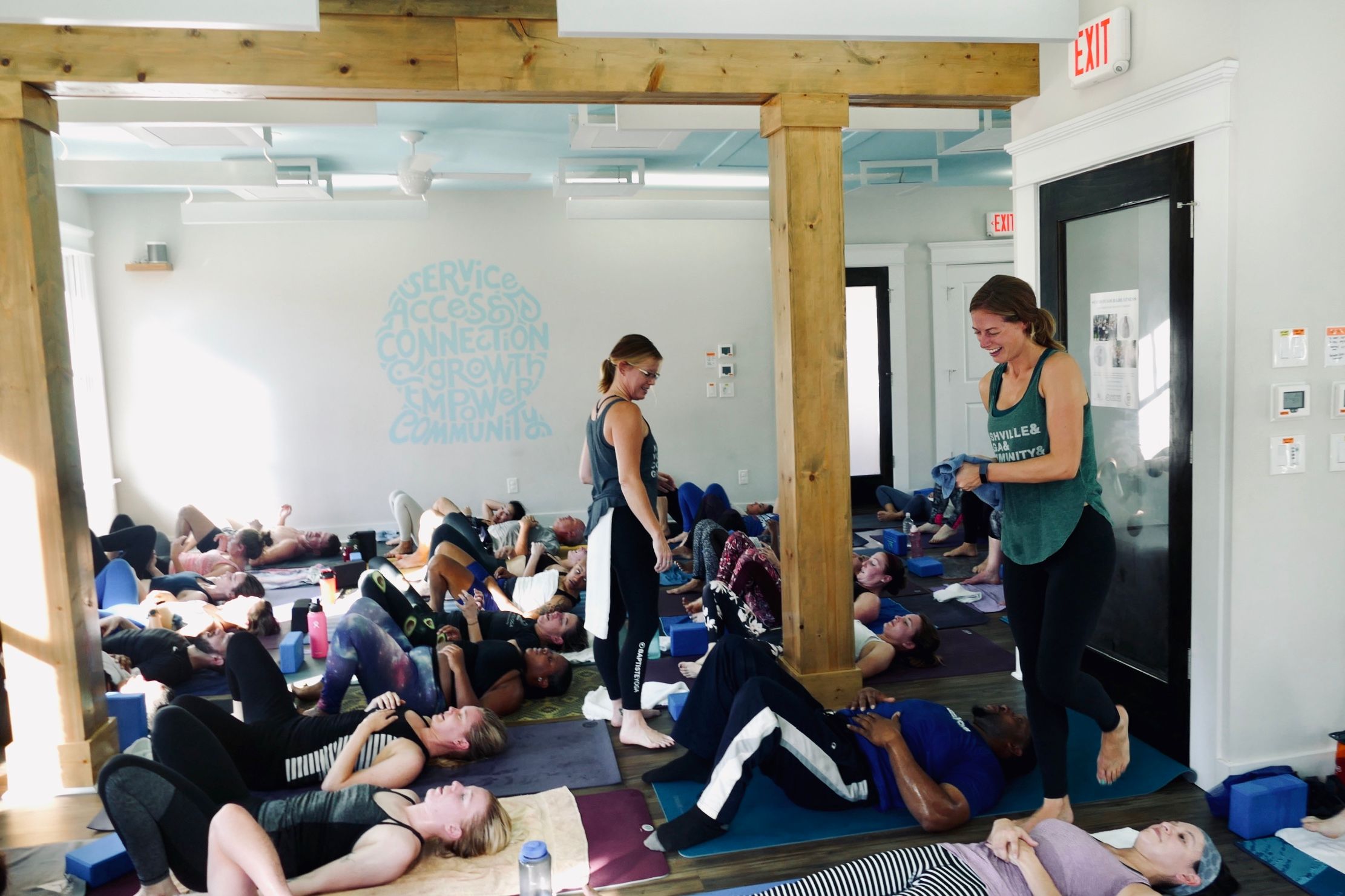 Two yoga teachers smile and walk through a studio of students lying on their backs